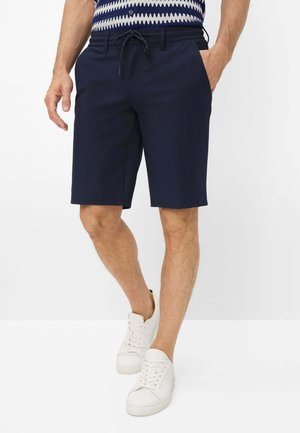 Man draagt marineblauwe shorts met trekkoord, witte sneakers en een gebreide top met blauw-wit patroon, staand met handen in de zakken.