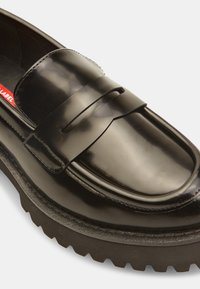 Mocassino in vernice nera con punta rotonda, dettagli del cinturino in evidenza e suola in gomma spessa con motivo zigzag.