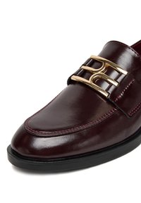 Burgundi bőrből készült papucs fényes felülettel, arany csattal és kontrasztos piros varrással a varrás mentén. Lekerekített orrú kialakítás.