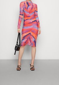 Tajtsittande långärmad klänning med ett gradient av orange, lila och blått. Har ruchérad detaljering och en böjd fåll. Matchad med svarta sandaler.
