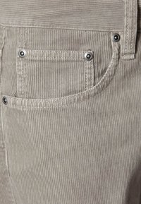 Beżowa tkanina sztruksowa z pionowymi żebrowaniami, wyposażona w małą kieszonkę z srebrnymi metalowymi zatrzaskami i subtelnymi detalami szwów.