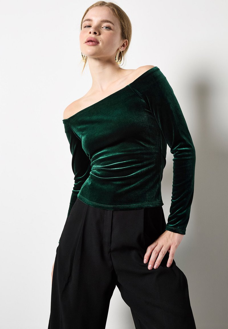 Grünes Samt-Oberteil mit schulterfreiem Design, langen Ärmeln und taillierter Passform. Glatte Textur, subtiler Glanz und geraffte Details an der Taille.