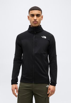 Jeune homme aux cheveux courts portant une veste noire zippée The North Face et un pantalon vert olive The North Face, debout devant un fond uni.