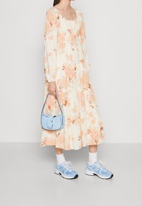 Kwiatowa midi sukienka w kremowym kolorze z pomarańczowym i różowym wzorem, długie rękawy, warstwowy fason; zestawiona z jasnoniebieskimi trampkami i małą niebieską torebką.