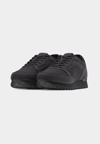 Paire de baskets basses noires en tissu texturé, lacets à l'avant et semelles épaisses en caoutchouc, inclinées sur fond blanc.