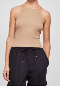 Žena ve fitted béžovém ribbovaném topu s tenkými ramínky a volných černých kalhotách se stahovací šňůrkou, ruce v kapsách, neutrální pozadí.
