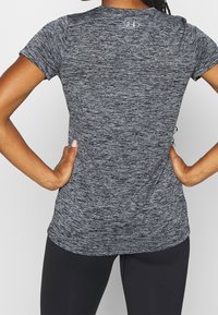 Žena v krátkém sportovním tričku šedé melírované barvy a černých legínách, s rukama v bok, zobrazena zezadu na jednobarevném bílém pozadí.