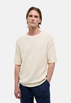 Jeune homme aux cheveux bruns mi-longs porte un pull crème texturé à manches courtes et un pantalon bleu foncé, debout avec une main dans la poche.