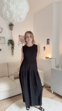 Vestido maxi preto sem mangas com um design em camadas, tecido texturizado e decote redondo. Usado com sandálias pretas de slide, ambientado numa sala iluminada.