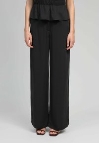 Femme portant un pantalon large noir et des sandales carrées bordeaux, se tenant contre un fond gris clair uni.