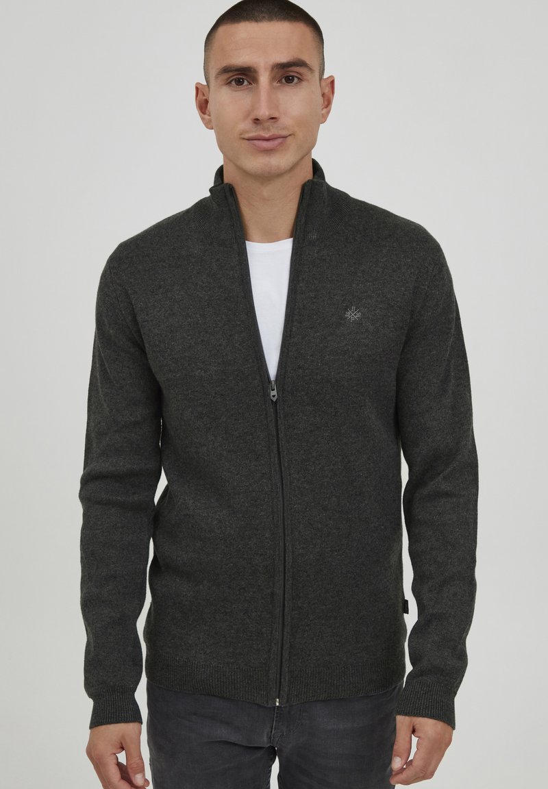 Cardigan zippé gris foncé en tissu tricoté doux, avec un col montant et des poignets côtelés, arborant un petit logo sur la poitrine.