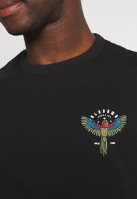 Felpa nera con colletto a coste presenta un colorato design ricamato di un pappagallo e la scritta "ALABAMA" e "WILD FREE" sotto.