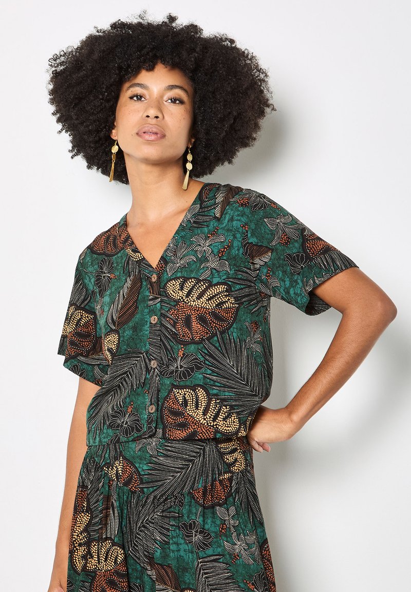 Femme aux cheveux bouclés portant un haut vert à boutons et une jupe assortie à grands imprimés de feuilles tropicales, posant avec une main sur la hanche.
