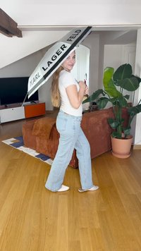 Črno-bela dežna palma Karl Lagerfeld z izrazitim besedilom, ki jo drži oseba v belem majici in svetlih modrih kavbojkah, v notranjem prostoru.