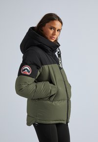 Doudoune avec un haut noir et un bas vert olive, dotée d'une grande capuche, d'un zip frontal et d'un patch logo rond sur la manche.