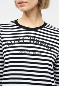 Κοντινό πλάνο ενός ατόμου που φοράει μαύρο και άσπρο ριγέ πουκάμισο με την επιγραφή «SAINT TROPEZ» και «MICHAEL KORS» στο μπροστινό μέρος.