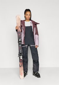 Conjunto de ski com uma jaqueta em bordeaux e lilás claro, calças overall pretas e botas de ski pretas. A segurar um ski com uma base castanho claro e fixações.