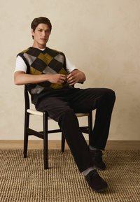 Grüner, schwarzer und grauer Argyle-Pullover über einem weißen Hemd; dunkle Cordhosen; dunkle Schuhe; sitzt auf einem Holzstuhl; gewebter Teppich darunter.