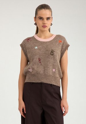 Jeune femme aux cheveux tirés en arrière porte un pull sans manches marron avec des étoiles, un cœur, une rose et un cygne brodés, ainsi qu’un pantalon sombre taille haute.