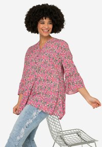 Blouse rose à motifs floraux avec un col en V, des manches trois-quarts avec volants, et une coupe ample et fluide, associée à un jean clair usé.