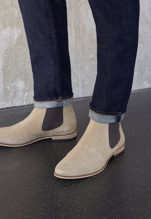 Hellbeige Wildleder-Chelsea-Stiefel mit dunklen elastischen Seitenteilen, getragen zu dunklen hochgekrempelten Jeans auf einem schwarzen gesprenkelten Boden.