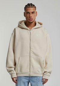 Felpa con zip colore beige realizzata in tessuto morbido, dotata di una grande tasca anteriore e di un logo ricamato sul lato sinistro del petto. Include tasche a marsupio.