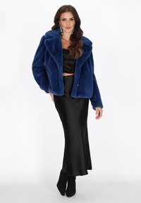 Kvinde iført en blå faux fur jakke over et sort satin sæt, stående med den ene hånd i lommen og sorte hæle støvler.