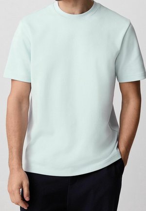Homme portant un t-shirt simple bleu clair à manches courtes avec col rond, et un pantalon noir, une main dans la poche.