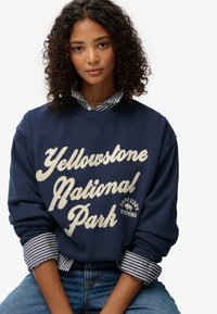 Sudadera azul marino con el texto blanco "Parque Nacional Yellowstone", con cuello redondo y puños a rayas. Combinada con jeans azules.