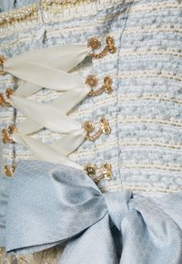 Tessuto azzurro chiaro texturizzato con fili d'oro, caratterizzato da lacci in satin bianco e ganci decorativi. Include un fiocco abbinate con un motivo geometrico.