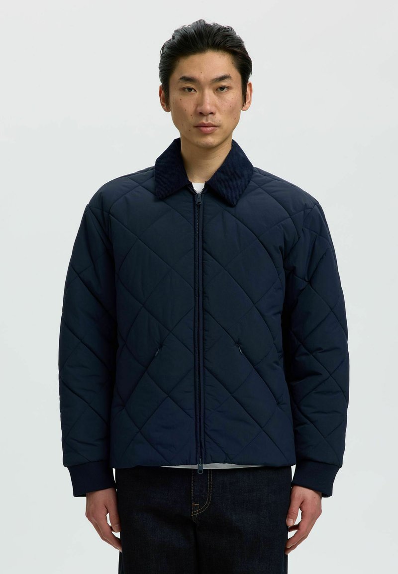 Homme portant une veste matelassée bleu foncé avec un col en velours côtelé, une fermeture éclair à l'avant et des poches, debout devant un fond clair uni.