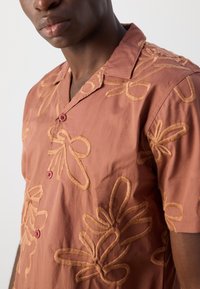 Camicia a maniche corte in tessuto terracotta con motivi floreali ricamati in beige, colletto con tacche e due bottoni rossi.