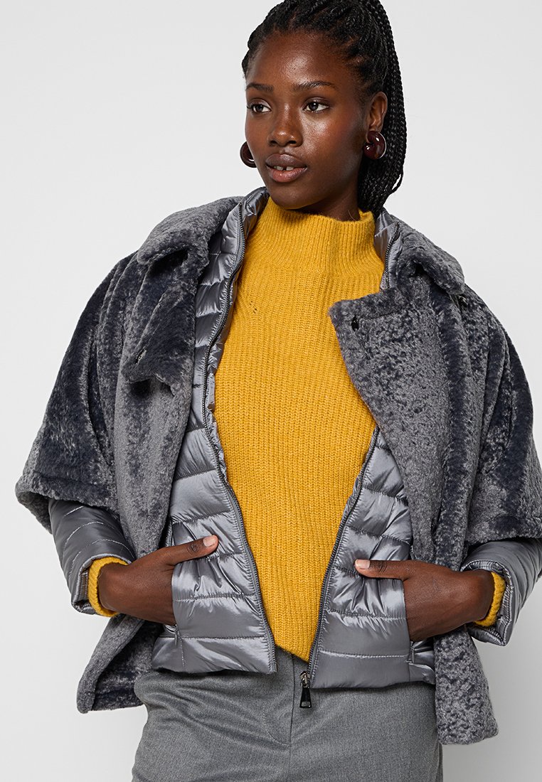 Graue Daunenjacke mit weichem, plüschigem Außenmaterial, ausgestattet mit einem hohen Kragen und Druckknopfverschluss, kombiniert mit einem leuchtend gelben, gestrickten Pullover.
