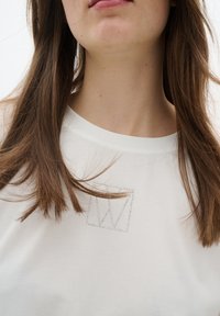 Valge T-särk ümmarguse kaeluse ja lühikeste varrukatega, mille esiküljel on hõbedane sädeleva rhinestone'iga ruudukujuline grafika. Sile tekstuur.