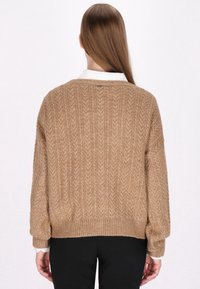 Pulover din tricot cu model de cablu, de culoare maroniu deschis, cu o croială lejeră, marginile și mansetele cu model cordon; purtat peste o cămașă albă cu guler, punând în evidență un model texturat.