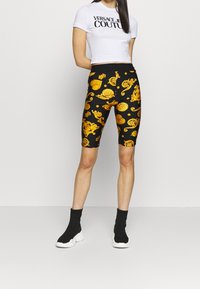 Black cycling shorts featuring a gold ornate pattern, paired with a white cropped t-shirt displaying black text. Black high-top sneakers.