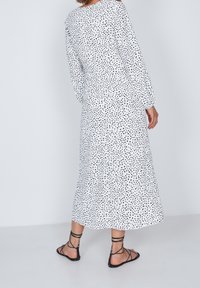Vestido blanco de manga larga con lunares negros, tejido fluido y corte relajado. Combinado con sandalias negras de tiras, vista lateral.