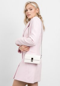 Borsa bianca con una finitura testurizzata, forma rettangolare e tracolla in catena argentata. Presenta una chiusura metallica ed è indossata con un blazer rosa pastello.