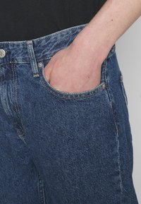 Blå denimjeans med en avslappnad passform, som har en framficka och synliga sömmar. Tyget har en klassisk blekning som framhäver dess textur.