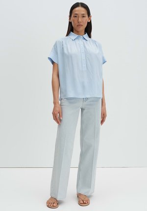 Femme debout devant un fond uni, portant un chemisier à manches courtes rayé bleu clair, un jean droit délavé et des sandales couleur sable.