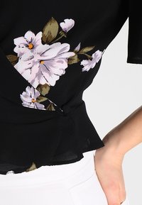 Schwarze Bluse mit hellrosa und weißen Blumenmotiven sowie grünen Akzenten, aus leichtem Stoff, mit Wickeldesign und Rüschensaum.