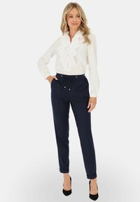 Kobieta w białej bluzce z falbanami i granatowych spodniach na sznurki, w czarnych szpilkach, stojąca z jedną ręką w kieszeni, spoglądająca w bok.
