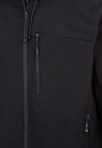 Czarna kurtka z bliska, pokazująca dwie pionowe kieszenie na zamek błyskawiczny na piersiach oraz centralny zamek z sznurkami na miękkim materiale.