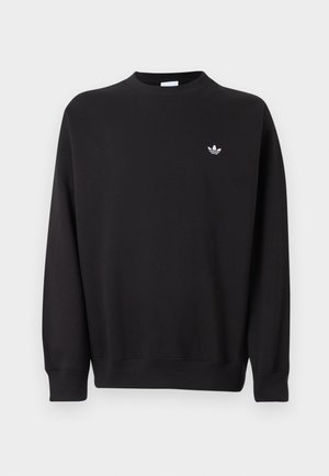 Mann mit kurzen lockigen Haaren trägt schwarzen Adidas-Sweatshirt und schwarze Jogginghose mit weißen Streifen, steht vor einem schlichten hellen Hintergrund.