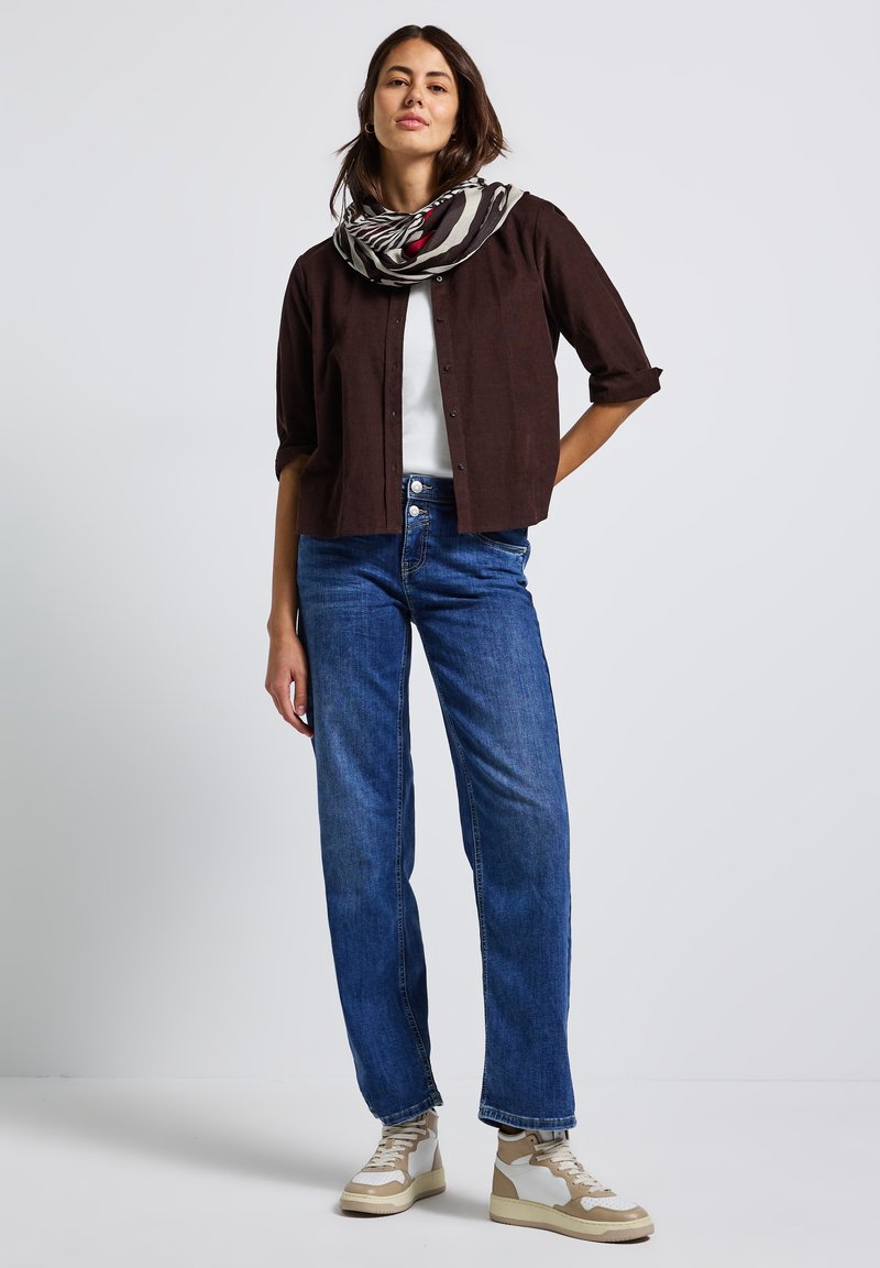 Donna in piedi che indossa una camicia button-up marrone scuro, una maglietta bianca, jeans blu, scarpe beige e una sciarpa con un motivo bianco e nero.