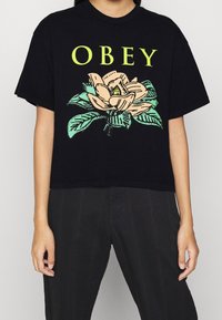 Černé zkrácené tričko s květinovým designem v béžové a zelené barvě. Text "OBEY" jasně žlutý nad květem magnolie a listy.