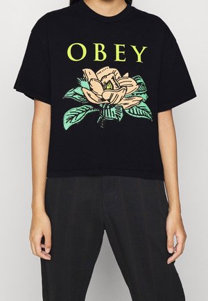 Black cropped t-shirt with a floral design in beige and green. "OBEY" text in bright yellow above a magnolia flower and leaves.