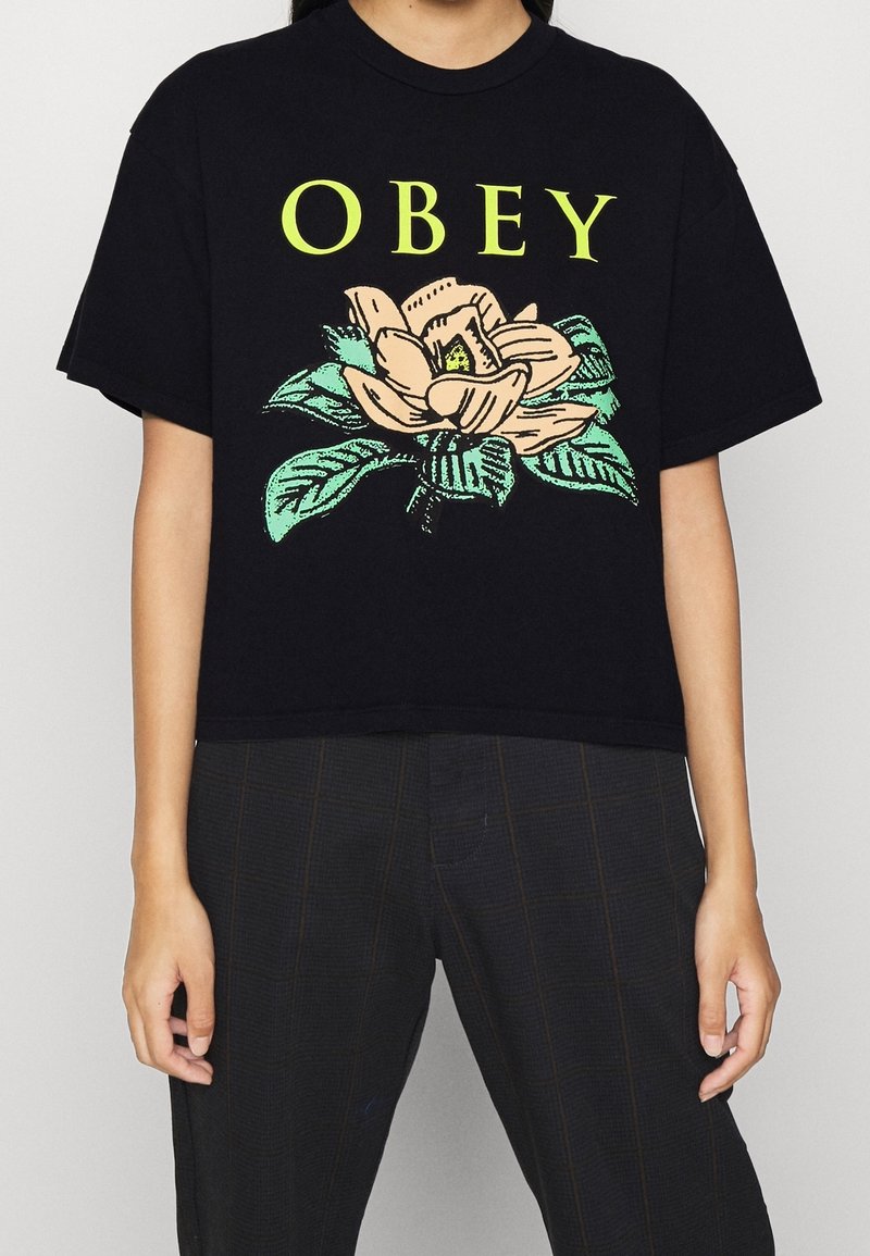 Černé zkrácené tričko s květinovým designem v béžové a zelené barvě. Text "OBEY" jasně žlutý nad květem magnolie a listy.