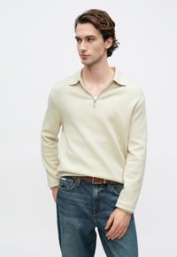 Kremowa bluza na zamek z kołnierzem, wykonana z teksturowanej tkaniny, zestawiona z niebieskimi dżinsami i brązowym paskiem.