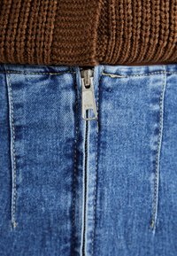 Visoko pasirana modra kavbojka s srebrnim YKK zadrgo spredaj, z diskretnim šivom in rahlo zbledelim izgledom tkanine.